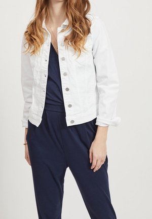 Woman wearing white buttoned denim jacket over navy blue V-neck jumpsuit, standing against a plain light background.