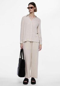 Camisa de botón de color beige claro con mangas largas, combinada con pantalones de pierna ancha a juego. Sandalias de plataforma negras y un bolso tote negro texturizado.