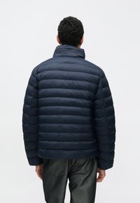 Geacă matlasată de culoare albastru închis, cu secțiuni matlasate orizontal, guler ridicat și manșete elastice. Confecționată din material neted și ușor.