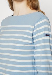 Pull-over à manches longues bleu clair avec des rayures horizontales blanches. Il présente un col rond et un patch logo sur la manche gauche.