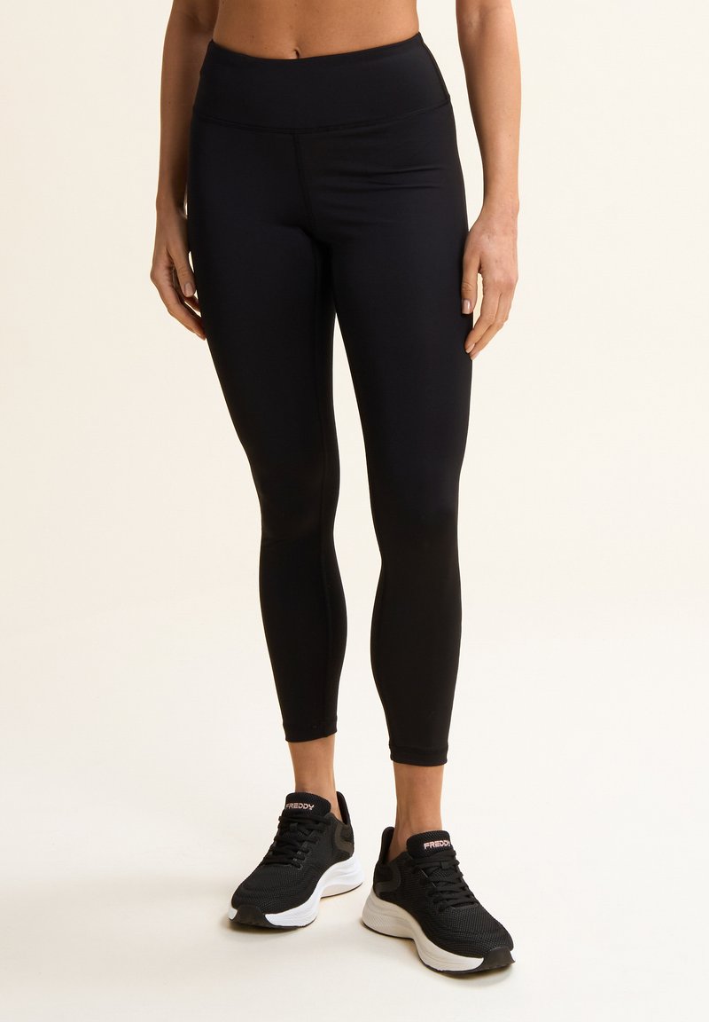 Leggings noirs taille haute en matériau extensible, avec une texture lisse et un design ajusté. Associés à des chaussures de sport noires.