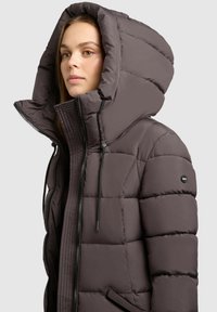 Puffer jakna u tamnosmeđoj boji s velikim kapuljačom. Ima kvadratni teksturirani uzorak, zatvaranje na zip i naglašavanje s vezicama na kapuljači. Lagani materijal.