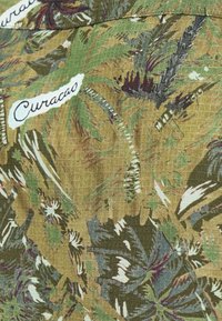 Πράσινο και καφετί υφασμάτινο με σχέδιο και υφή. Περιλαμβάνει φύλλα φοινίκων και τη λέξη "Κουρασάο" σε λευκό χρώμα σε φωτεινό φόντο.