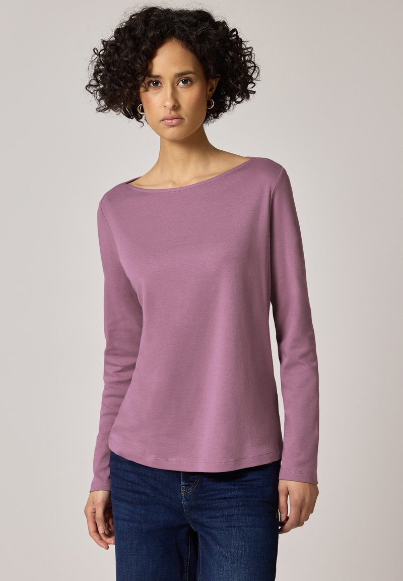 Mujer con cabello rizado que lleva pendientes de aro dorados, una camiseta de manga larga color malva y vaqueros azul oscuro, de pie frente a un fondo neutro.