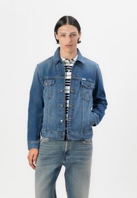 Ung mann står mot en hvit bakgrunn, iført en blå denimjakke over en stripete skjorte og lyse jeans, med et nøytralt uttrykk.