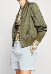 Olivgrön bomberjacka med ribbade muddars, dragkedjefickor och framficka, använd över en tie-dye topp och ljusblå shorts.