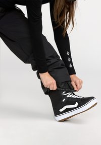 Sorte snowboarding støvler med hvide detaljer og snørebånd. Personen er ved at justere fodtøjet, som har en struktureret overflade og gummisål.