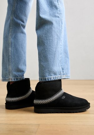 Botas de tobillo de ante negro con suela gruesa y una banda decorativa estampada en la parte superior. Llevadas con jeans de un azul claro en un entorno neutro.