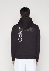 Zwarte hoodie met witte "Calvin Klein" logo op de achterkant. Gemaakt van zachte stof, met een ontspannen pasvorm en geribde manchetten.