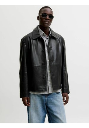 Mand iført sort læderjakke med knapper, ternet skjorte, blå jeans og solbriller stående foran en ensfarvet lys baggrund.