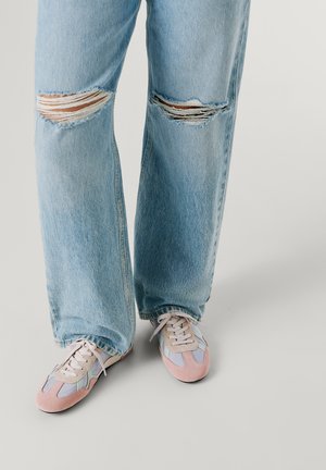 Világoskék szakadt farmer pasztellszínű sportcipővel, amely rózsaszín, bézs és kék részleteket tartalmaz, semleges háttéren.