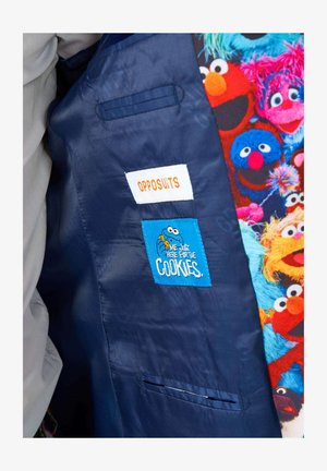 Laineesta valmistetun tummansinisen puku-takin sisäpuoli, jossa on valkoinen etiketti, jossa lukee "OPPOSUITS", sekä sininen patch, jossa on kuvitteellinen keksi-hahmo ja teksti.