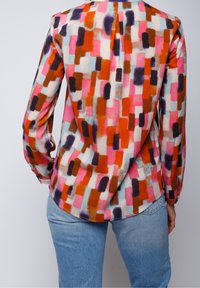 Blusa multicolore con motivi astratti in rosso, rosa, arancione e marrone, maniche lunghe e un sottile dettaglio di piega sul retro.