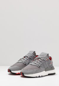 Šedé sportovní boty s texturovaným vrškem z mřížkované tkaniny, semišovými akcenty, červenými detaily a bílou polstrovanou podrážkou s černým vzorkem.