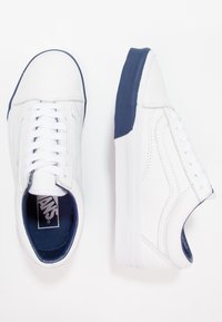 Sneakers bianchi realizzati in materiale sintetico con accenti blu navy, dotati di un design con lacci, suola testurizzata e loghi laterali.