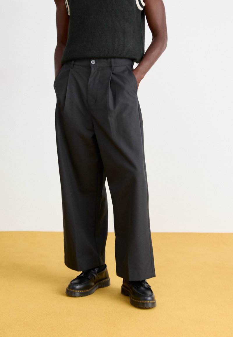 Black wide-leg trousers with front pleats and a button closure. Worn with black shoes, displayed against a yellow floor backdrop.
