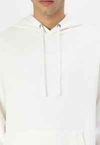 Hanorac alb dintr-un material moale, cu buzunar frontal, șnururi ajustabile și logo embosat. Design slim fit.