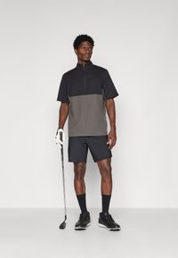 Πόλο γκολφ κοντομάνικο σε μαύρο και γκρι, με μπροστινό φερμουάρ, συνδυασμένο με μαύρα σορτς. Το μοντέλο κρατά ένα μπαστούνι γκολφ, φοράει μαύρα παπούτσια.