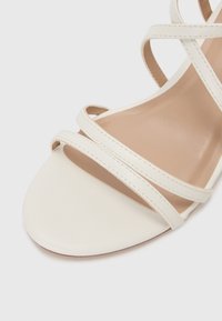 White leather sandal with thin crisscross straps, open toe, and a smooth, rounded sole. Stitching detail on straps adds texture.