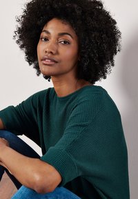 Grüner strukturierter Strickpullover mit weiten Ärmeln, getragen von einer Person, die sitzt und Jeans trägt. Glatter Hintergrund in neutralen Tönen.