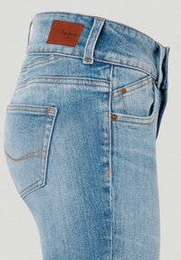 Jean en denim bleu clair avec un patch de marque en cuir marron sur la ceinture, poches visibles à l'avant et à l'arrière, et surpiqûres contrastées.