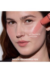 Blushstick in koraal aangebracht op de wang, gladde textuur op de huid. Subtiele gloed, model met een medium huidtint, minimale make-up, neutrale lippen.
