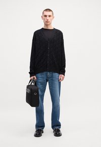 Mustaa kuviollista neuletakkia, jossa on V-pääntie ja napit, yhdistettynä sinisiin farkkuihin ja mustiin nahkakengiin. Mallilla on musta laukku.