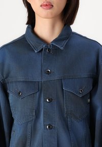 Τζάκετ από τζιν σε σκούρο μπλε χρώμα με κλασικό γιακά, δύο τσέπες στο στήθος και μαύρους στύλους; διαθέτει ορατές ραφές και λεία υφή.