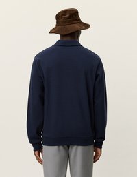 Tumši zils džemperis ar apkakli, ribināti apkakles un piedurkņu gali. Valkāts ar brūnu korduroja vāciņu. Vienkārša auduma, ikdienišķs dizains.