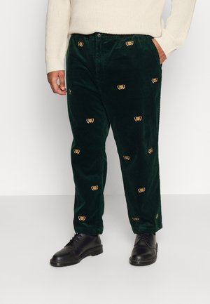 Mand iført mørkegrønne kordfløjlsbukser med broderede tigeransigter, offwhite sweater og sorte snørestøvler, står med den ene hånd i lommen.