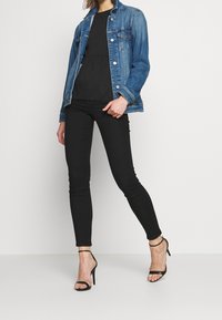 Femme portant une veste en denim bleue, un chemisier noir, un pantalon skinny noir et des sandales à talons hauts noires, debout devant un fond blanc.