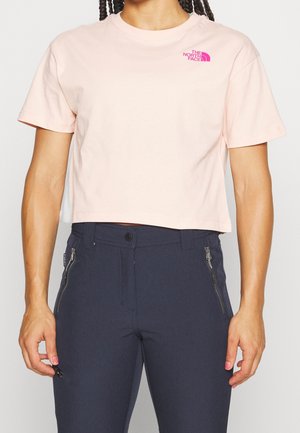 Kobieta ubrana w krótki, jasnoróżowy t-shirt marki The North Face oraz ciemnogranatowe spodnie z kieszeniami na zamek, stojąca z ramionami swobodnie opuszczonymi.