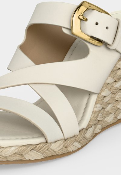 Sandale compensée blanche avec brides croisées en cuir, boucle dorée et détail de semelle en jute tressée.