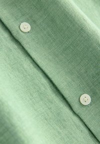 Tissu en lin vert clair avec une finition texturée ; présente un bouton blanc à quatre trous, mettant en avant une apparence lisse et douce.