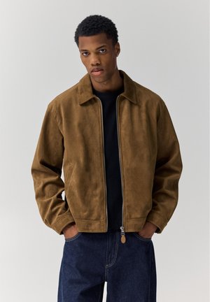 Tânăr îmbrăcat în geacă maro din piele întoarsă cu fermoar peste un tricou negru, cu mâinile în buzunare, pe un fundal simplu și deschis la culoare.