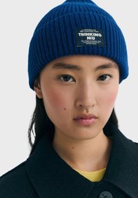 Joven mujer con un gorro de lana azul con una etiqueta de "Thinking Mu", abrigo negro y top amarillo, contra un fondo claro y uniforme.