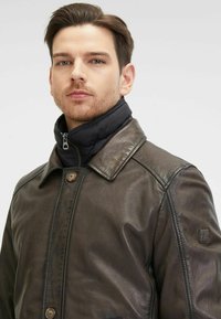 Braune Lederjacke mit Kragen, ausgestattet mit einem schwarzen, mit Reißverschluss versehenen Innenkragen. Besondere Details sind zwei vordere Knöpfe und ein seitlicher Logo-Patch.