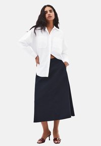 Weißes, oversized Hemd mit Knopfleiste und zwei Fronttaschen, kombiniert mit einem dunkelblauen, knielangen Rock und burgunderfarbenen, hochhackigen Sandalen.