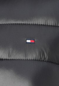 Μαύρο επενδεδυμένο ύφασμα με γυαλιστερή υφή, που διαθέτει ένα μικρό κεντημένο λογότυπο με οριζόντιες ρίγες σε κόκκινο, λευκό και μπλε.