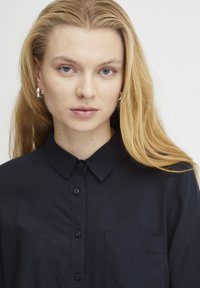 Camicia nera con bottoni, colletto, con tasca frontale, texture liscia e vestibilità comoda. Il modello ha i capelli lunghi e accessori minimali.