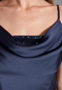 Abito di satin con scollatura a cowl. Colore blu scuro, con un accento di paillettes sullo scollo. Texture liscia, silhouette elegante.