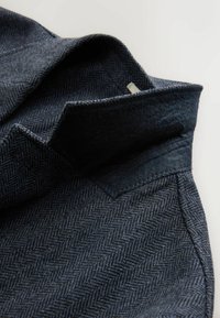 Marineblaues Fischgrät-Wollgewebe mit einer weichen, strukturierten Oberfläche. Der Kragen ist im Spitzstil mit kontrastierenden dunkleren Stoffkanten.