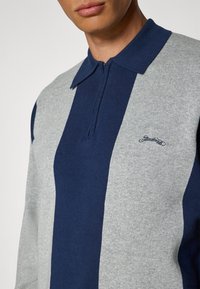 Marineblå og grå stripete polo med glidelås i halsen, med en teksturert stoff og et lite brodert logo på brystet.