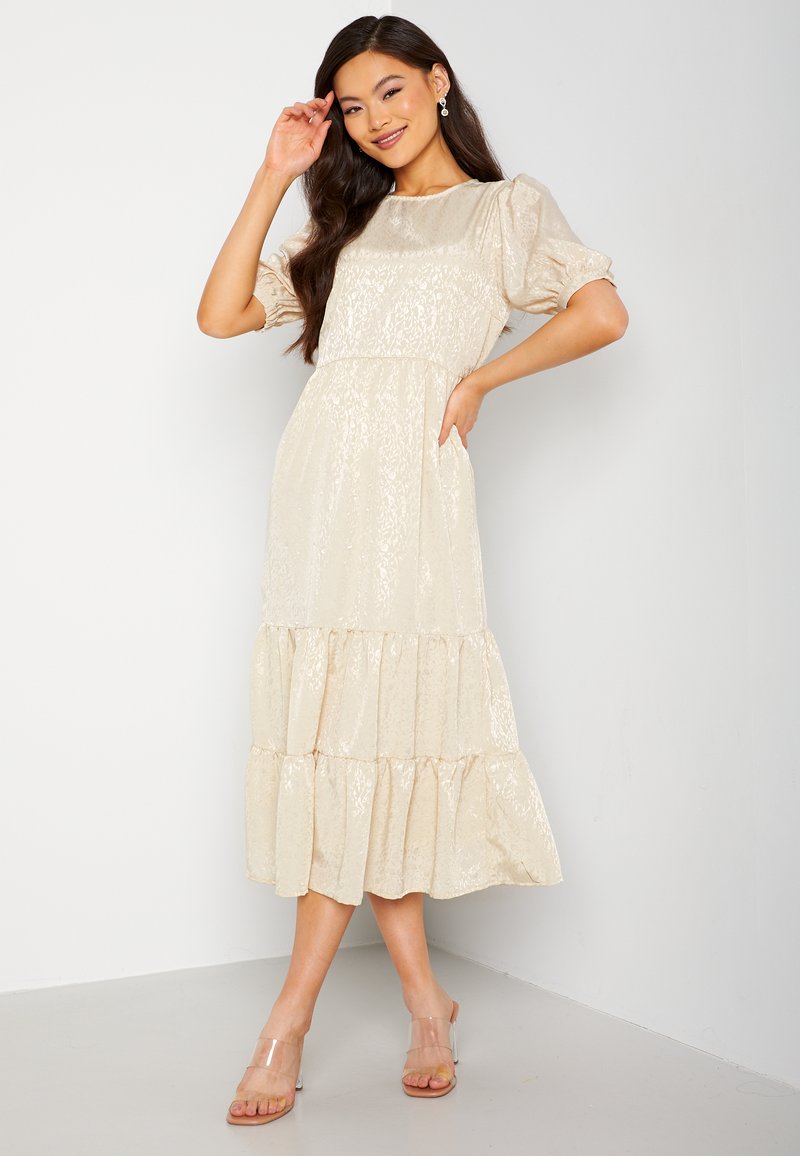 Beige, gelaagd maxi-jurk van een gestructureerde, glanzende stof, met pofmouwen en verborgen zijzakken. Gecombineerd met doorzichtige instap-sandalen.