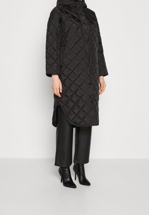 Personne portant un manteau matelassé noir d'une longueur jusqu'aux genoux avec un ourlet incurvé, un pantalon noir à jambes larges, et des bottes noires à talons hauts pointues.