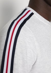 Sudadera gris con tres rayas paralelas en azul marino, blanco y rojo en el hombro, con un cuello redondo y textura suave.