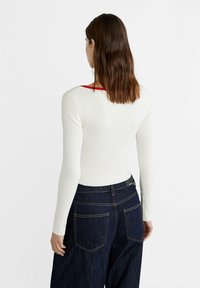 Witte geribbelde blouse met lange mouwen en een rode halslijn, gecombineerd met donkerblauwe wijde jeans. Achteraanzicht weergegeven, met nadruk op de pasvorm en textuur.