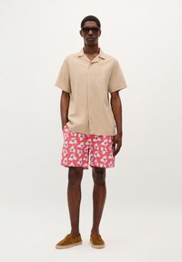 Light beige short-sleeve shirt, pink shorts with white and blue abstract pattern, brown espadrilles, and dark sunglasses worn by a male model.