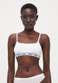 Balta medvilninė bralette su plonais dirželiais ir juodu logotipo juostu. Turi kvadratinį iškirpimą ir lygaus paviršiaus tekstūrą, dėvima su šviesios spalvos kelnaitėmis.