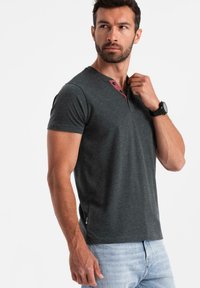 Mörkgrå kortärmad T-shirt med V-hals och två röda knappar. Mjuk textur, figurnära design och detalj med etikett i sidosömmen.